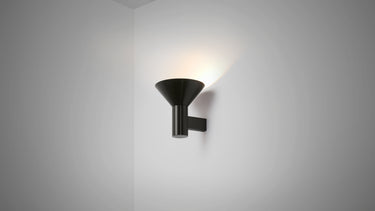 Torres wall light in bronze with a funnelled metal shade angled upward on a grey studio wall