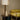 Roc table lamp with a natural linen square shade, antique bronze column and travertine block on a round side table beside a yellow chair