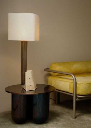 Roc table lamp with a natural linen square shade, antique bronze column and travertine block on a round side table beside a yellow chair