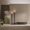 Two Torres floor lamps in bronze lit against a taupe wall beside a green armchair and a bench