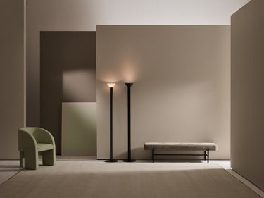Two Torres floor lamps in bronze lit against a taupe wall beside a green armchair and a bench