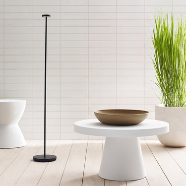 Moneta Medium floor lamp in Black beside a round white pedestal table with a bowl, tall grasses behind