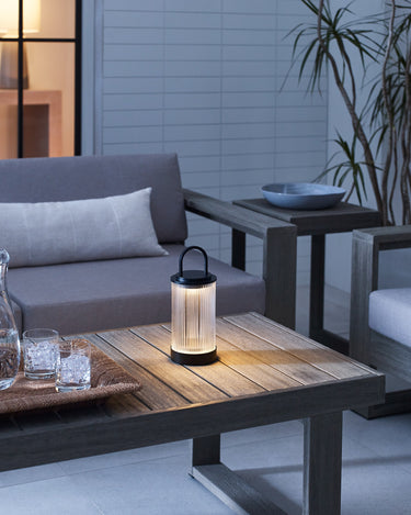 Tawa Accent rechargeable table lamp in Black, a ribbed glass lantern with carry handle, glowing on an outdoor table