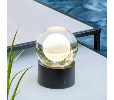 Black Mina Accent glass globe lamp glowing on a poolside table with grasses and water behind