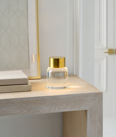 Caspe 7 rechargeable lamp in Hand-Rubbed Antique Brass glowing on a pale console beside a mirror and books