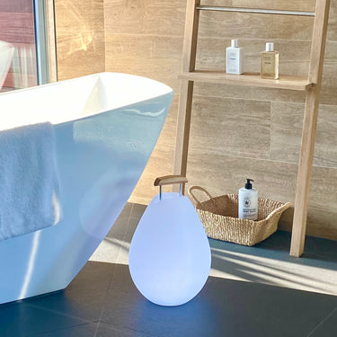 Vessel 2S Bluetooth LED Cordless Lamp by Smart & Green glowing soft blue on a bathroom floor beside a tub - LoftModern