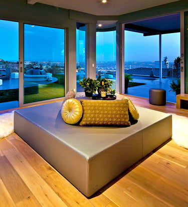 Play Pad square resort bed in gray vinyl with gold pillows and a tray, in a glass-walled living room at dusk