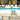 Two white Play Pad resort beds on a pool deck beside lounge chairs and an umbrella table
