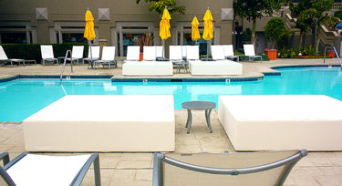 Two white Play Pad resort beds on a pool deck beside lounge chairs and an umbrella table