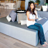 Woman reading on a gray Play Pad resort bed in a bright open lounge with wood floors