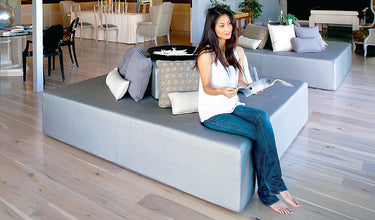 Woman reading on a gray Play Pad resort bed in a bright open lounge with wood floors