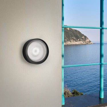 Oluce Berlin-Out black round wall lamp on a pale wall beside a window with a sea view