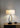 Stella table lamp with a linen drum shade on a wooden sideboard beside a small bowl against a grey wall