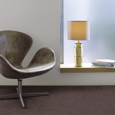 Ring table lamp in satin brass on a windowsill beside a grey leather armchair
