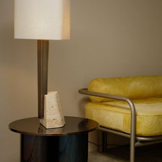 Roc table lamp with a natural linen square shade, antique bronze column and travertine block on a round side table beside a yellow chair