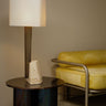 Roc table lamp with a natural linen square shade, antique bronze column and travertine block on a round side table beside a yellow chair