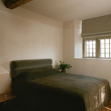 Cubist table lamp on a bedside table in a calm bedroom with a green velvet bed and a mullioned window