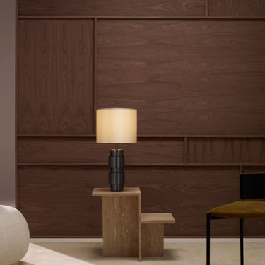 Ring table lamp in bronze with a lit silk shade on a wood stand against a walnut-paneled wall, beside a cream pouf