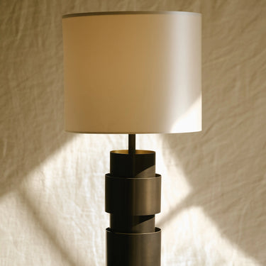Ring table lamp in bronze with a cream silk shade on a dark plinth, with shadows on the wall behind