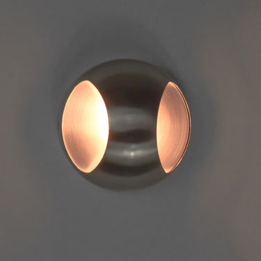 ET2 Wink wall sconce, Satin Nickel, lit with warm light glowing from both openings on a dark wall