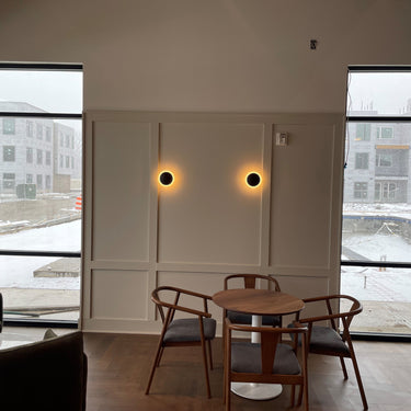 ET2 Saucer gold disc wall sconce, two glowing discs on a wall between tall windows in a lounge with a dining table