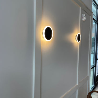 ET2 Saucer gold disc wall sconce, two glowing discs on a white paneled wall beside a window