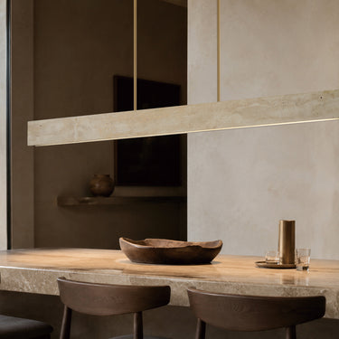 ET2 Lintel travertine linear pendant floating above a long wood dining table with brown chairs and a bowl, beige wall behind