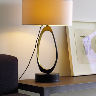 Stella table lamp glowing on a console beside a framed drawing and a dark sofa arm