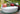 White Roll bolster along the back of a Moon Pad daybed with coral and green pillows beside a reflecting pond