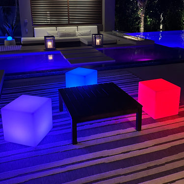 Violet, blue and red glowing Cube lamps around a dark coffee table beside a pool at night